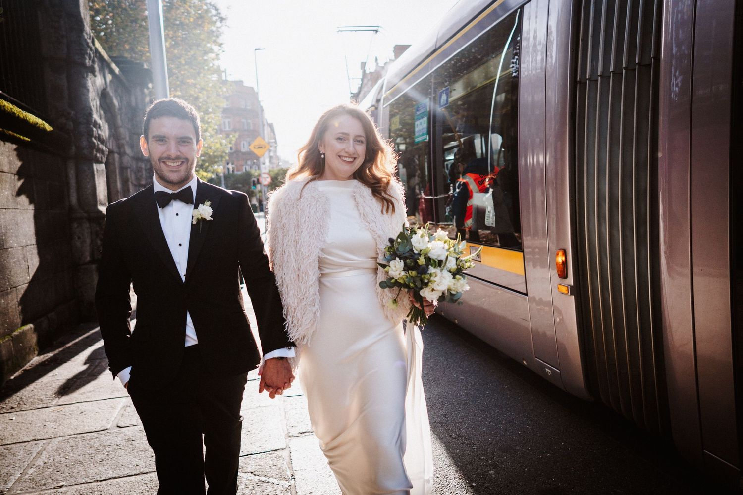 Wedding couple walks together through sunlit city street holding hands with tall buildings in background.