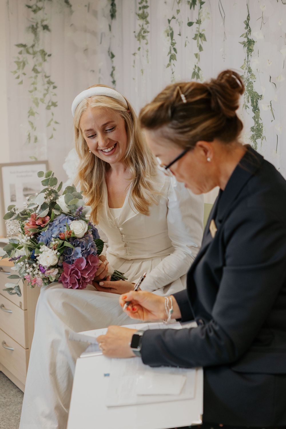 A radiant bride in white holds a colorful bouquet while signing documents with a registrar at a ceremony.