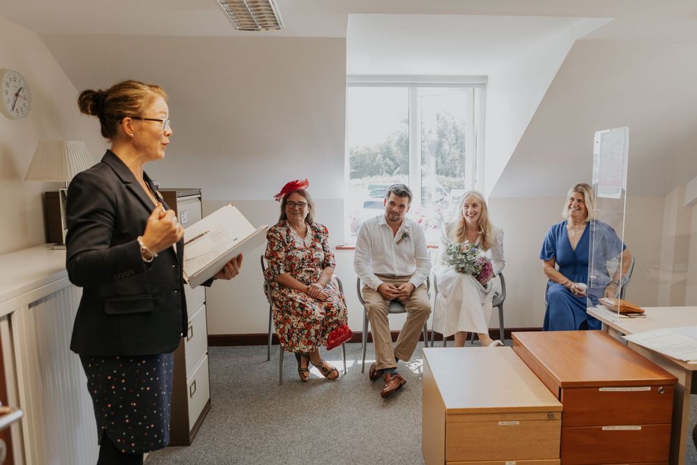 Wedding officiant reads ceremony script to small intimate group of guests seated in bright room.