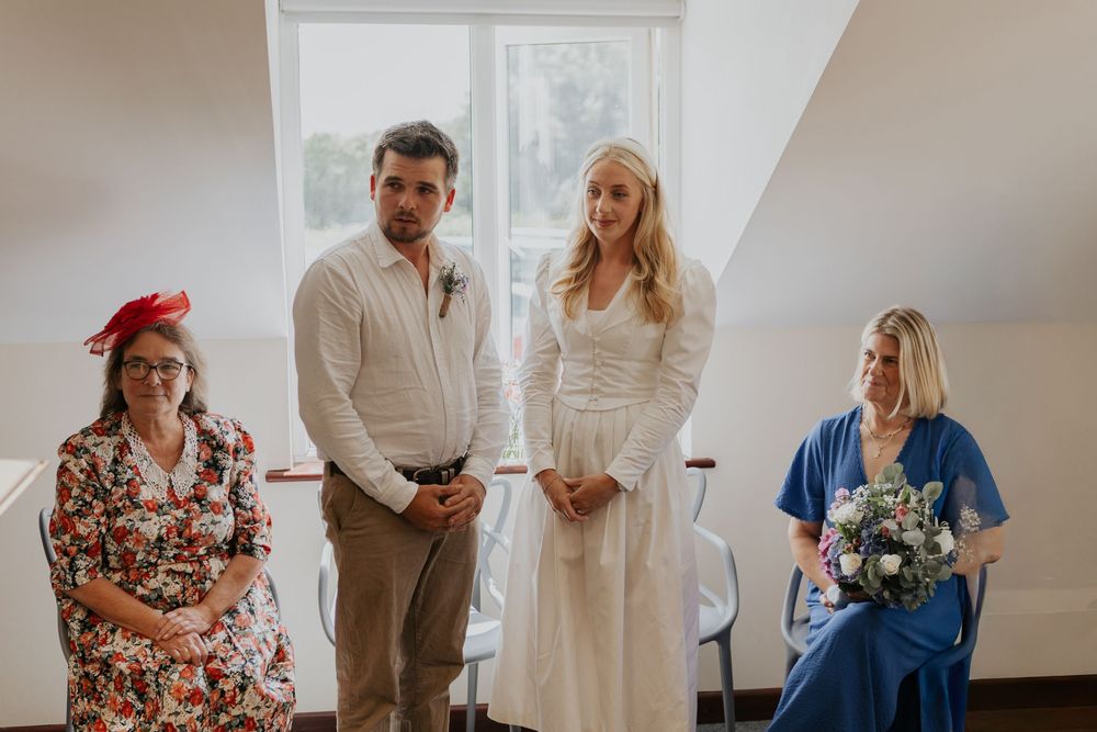 Bride in white dress and groom stand with two guests during small intimate civil wedding ceremony.
