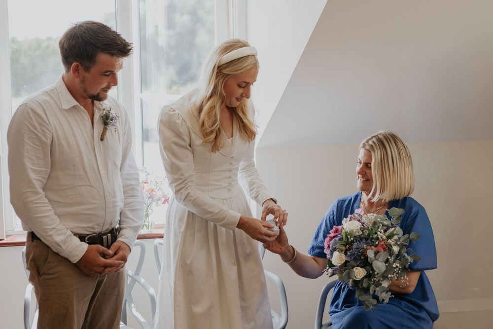 Wedding guest in blue dress hands ring box to smiling bride during intimate ceremony exchange.