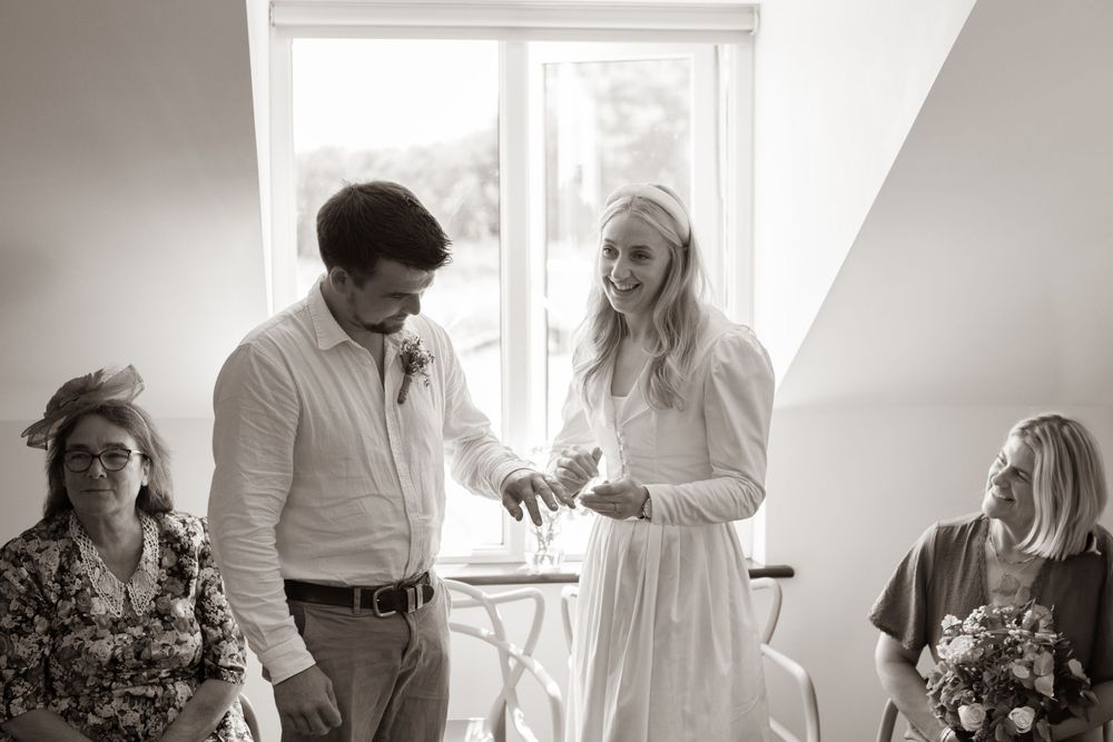 Bride laughs joyfully while exchanging wedding rings with groom in black and white ceremony photo.