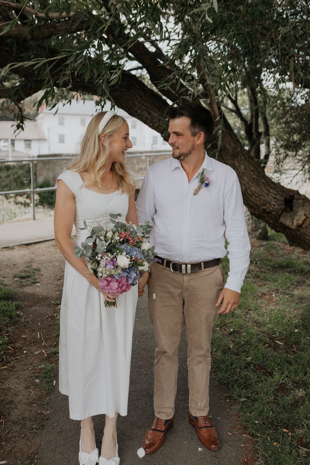 Bride holding colorful bouquet laughs with groom under large tree after outdoor wedding ceremony.