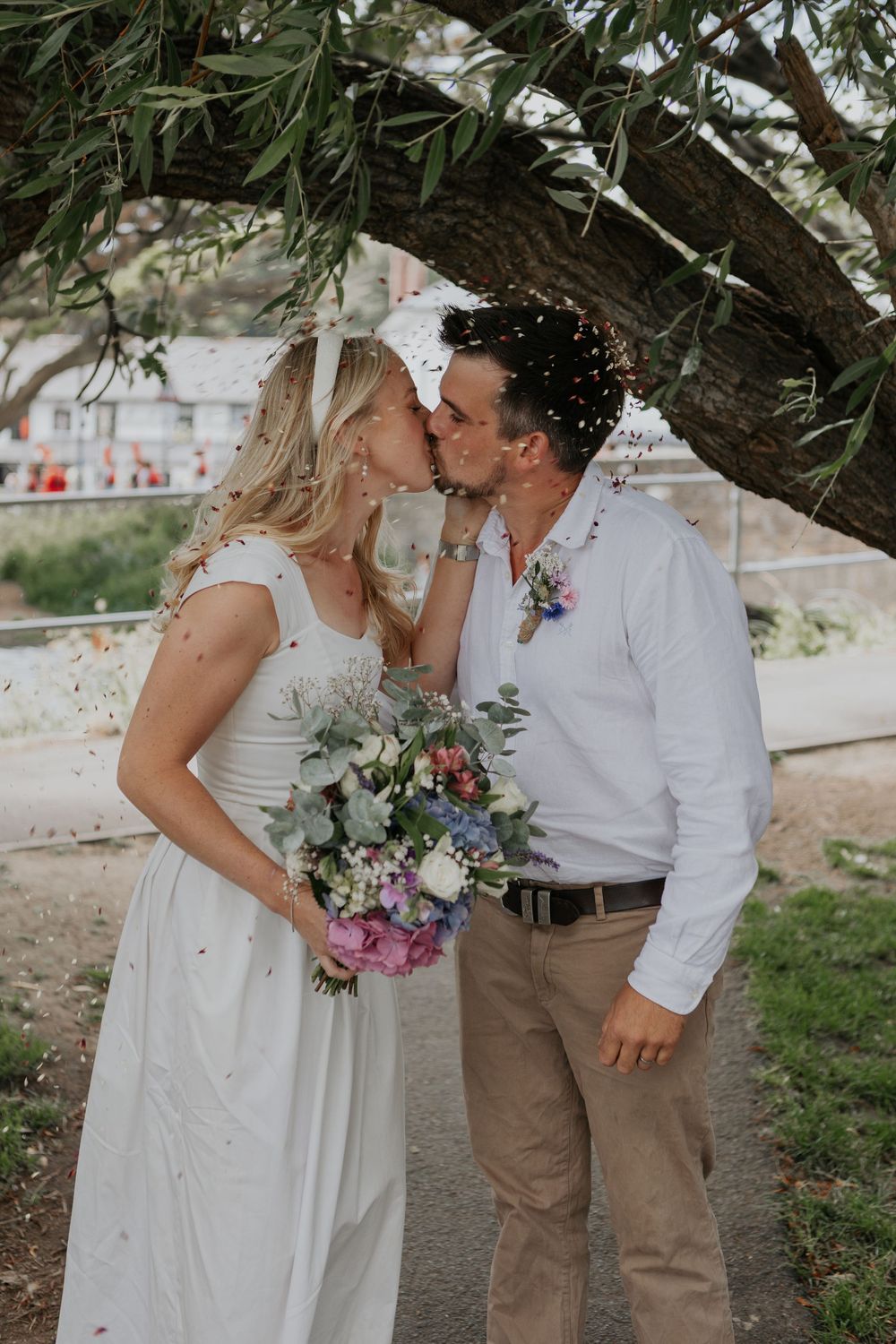 Newlyweds share first kiss under olive tree with colorful confetti falling during outdoor celebration.