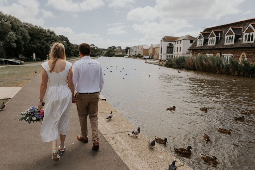 Bride and groom walking hand in hand along a riverside path with ducks, holding a colorful wedding bouquet.