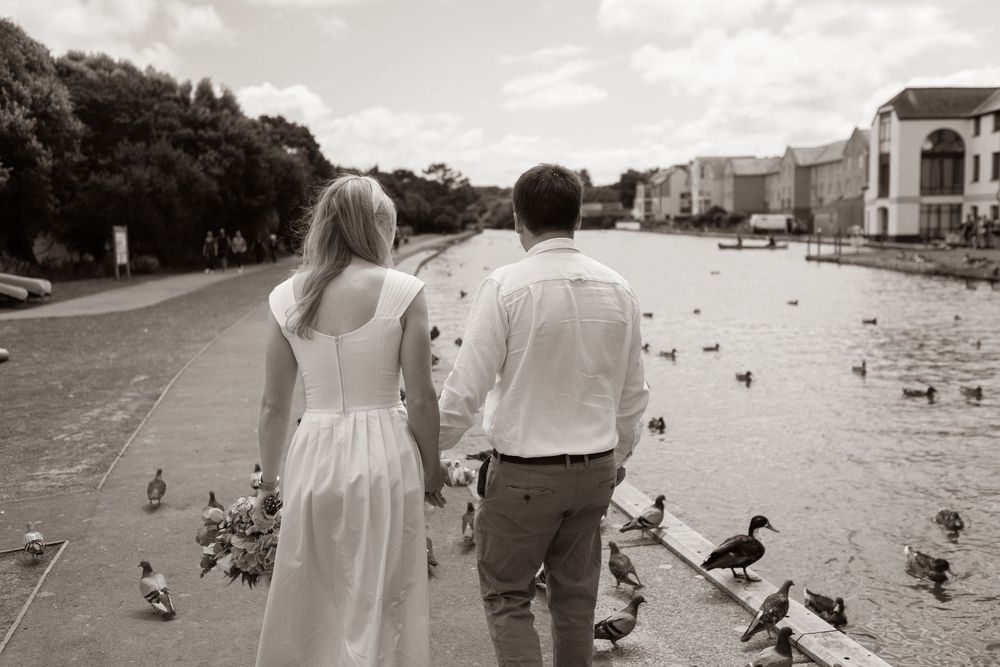 Newlyweds walk hand in hand along waterfront promenade with ducks in black and white wedding photo.