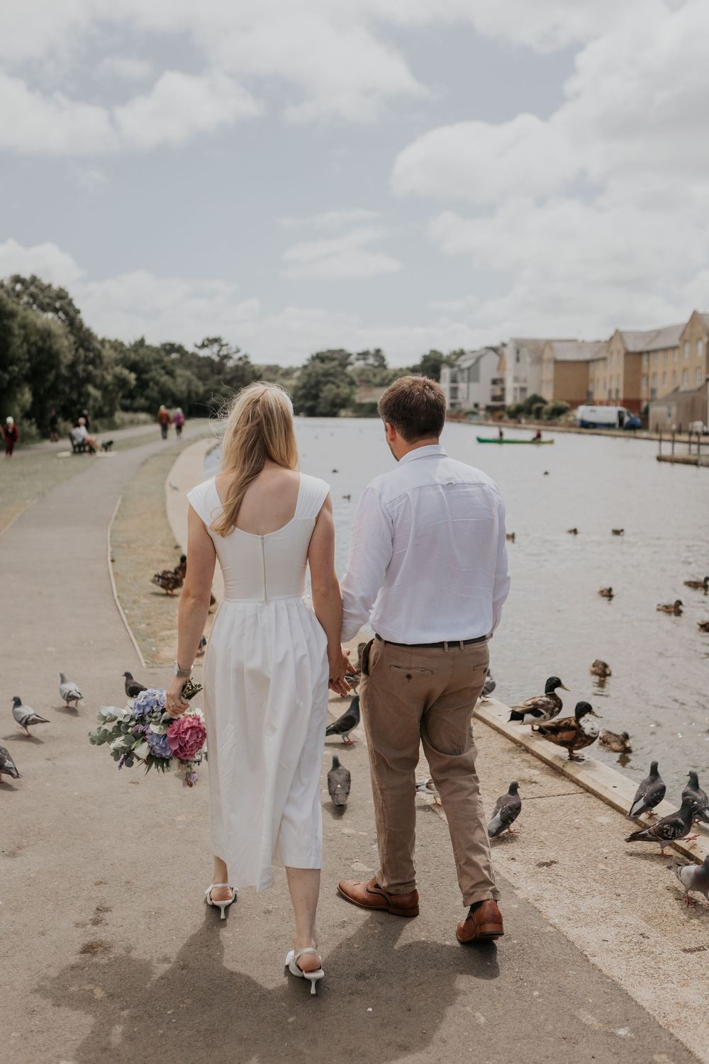 Newlywed couple walking by a duck-filled lake, bride in white dress holding a floral bouquet, groom in khakis.