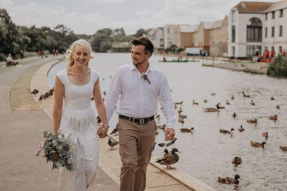 Smiling bride and groom holding hands by a scenic lake with ducks, bride carrying a blue and white bouquet.