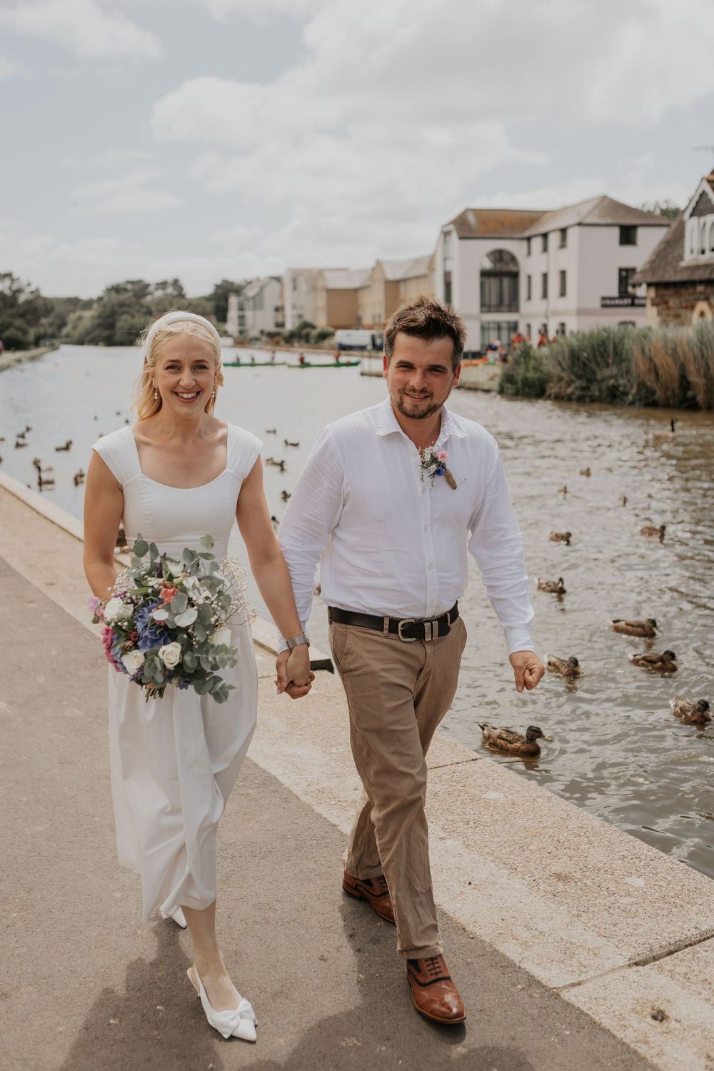 Happy bride and groom walking hand in hand along a waterfront path surrounded by ducks on their wedding day.