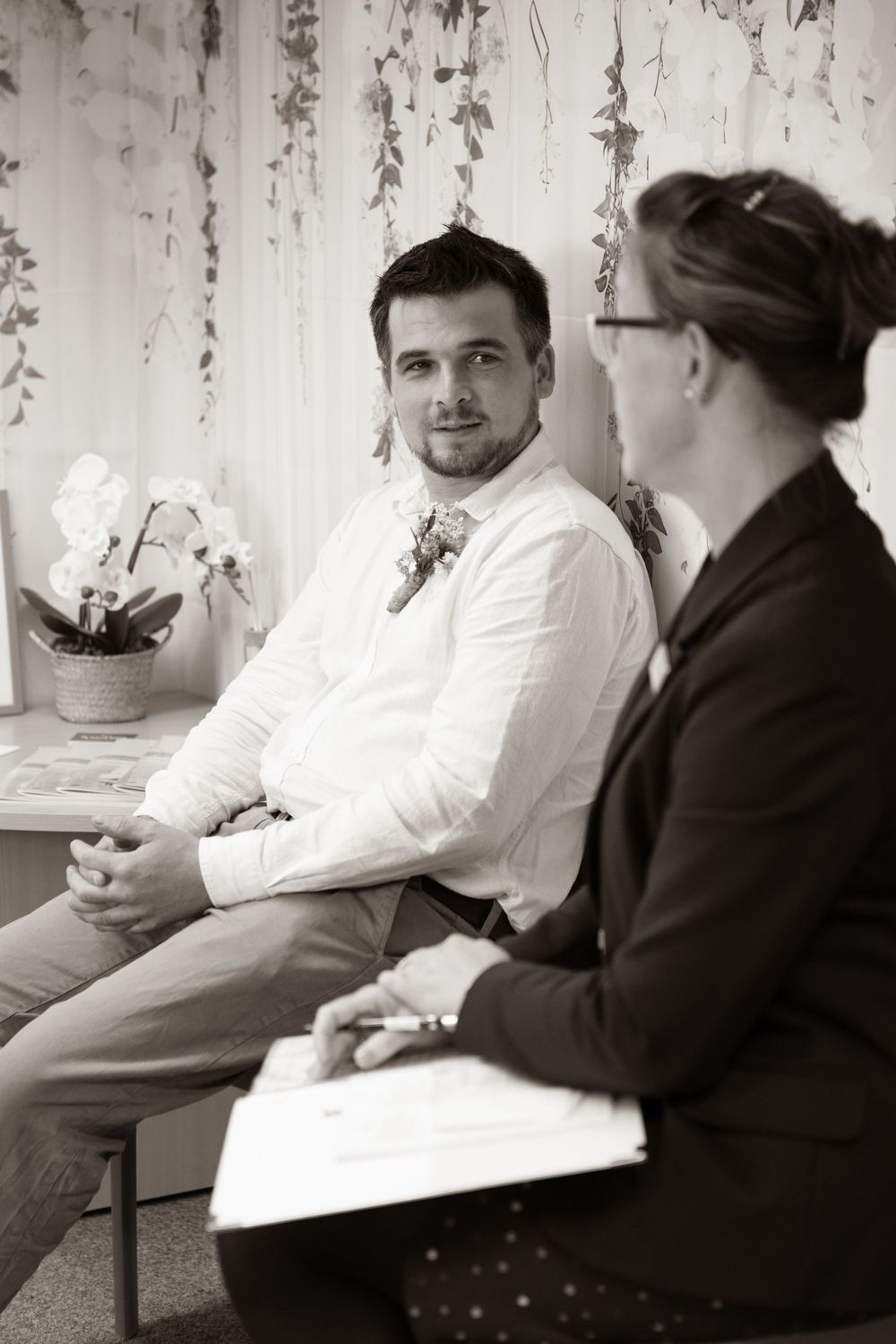 Groom in white shirt with boutonniere speaks with officiant before wedding ceremony in black and white photo.