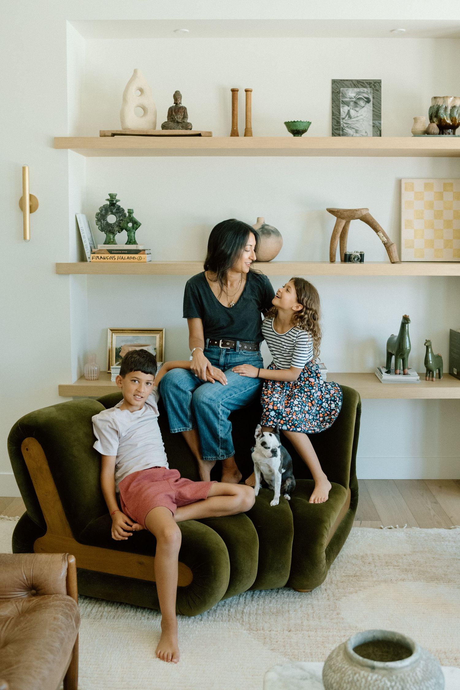 A family shares tender moments together on a dark green velvet couch with floating shelves displaying decor in the background.