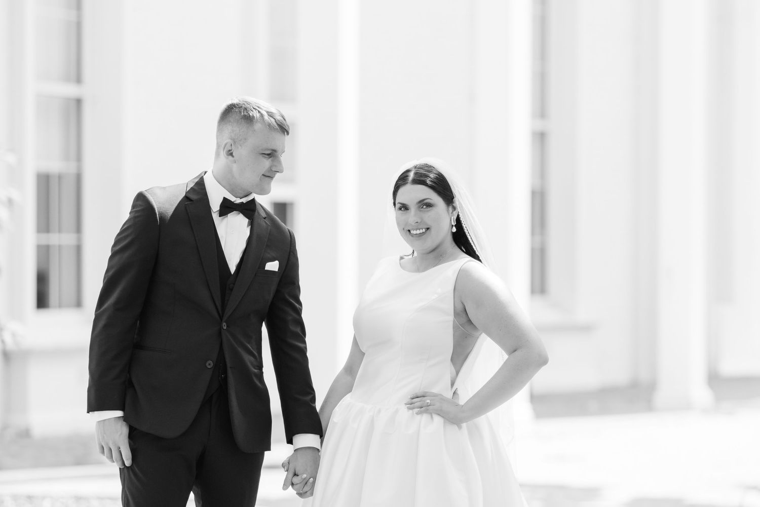 A bride and groom walk together in an elegant outdoor setting with white columns in the background.