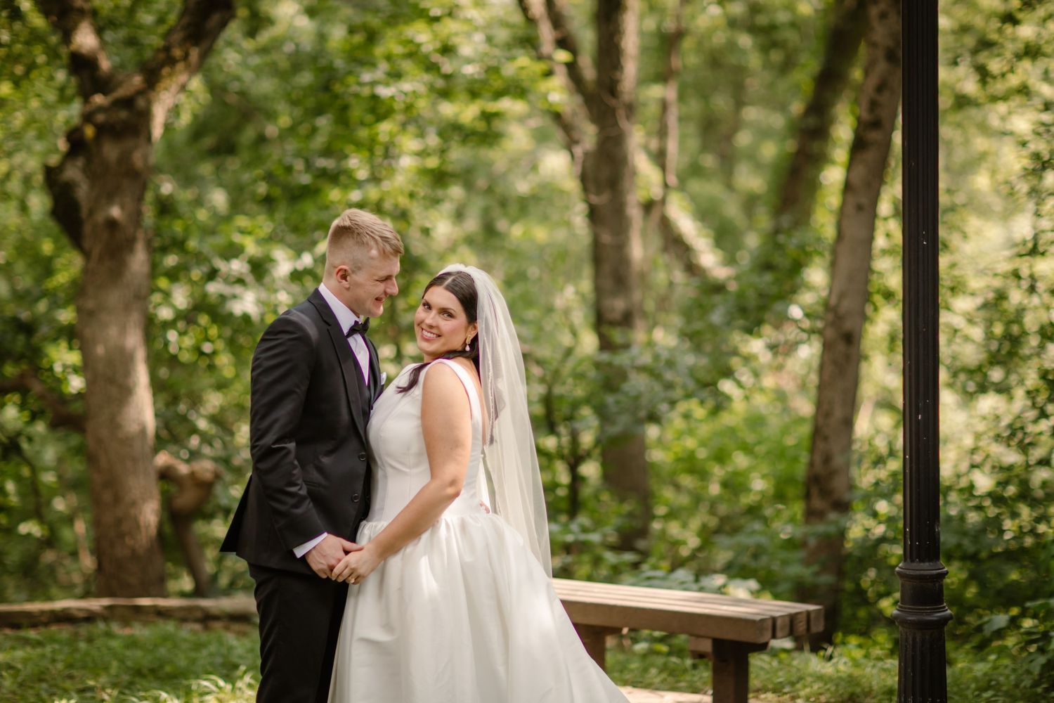 A bride and groom share a romantic moment together on a wooden bench surrounded by lush green trees in a forest setting.