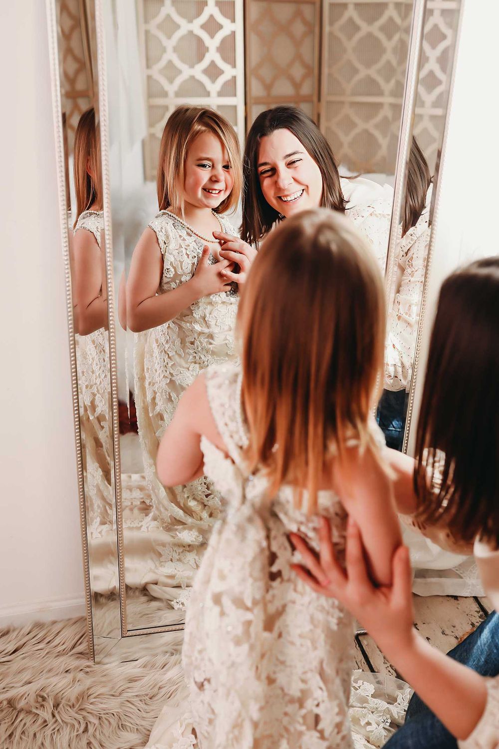 Two friends laughing and trying on elegant lace wedding dresses in front of ornate mirrors.