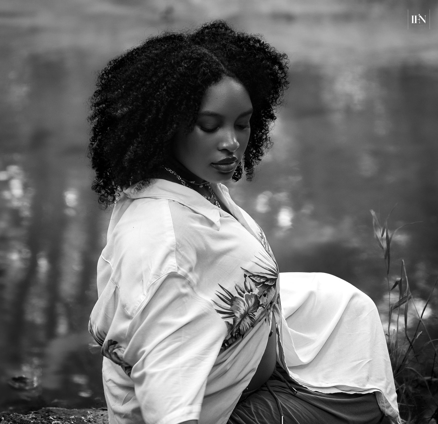 Black and white maternity portrait of a young woman with curly hair wearing a white shirt, sitting outdoors near water.
