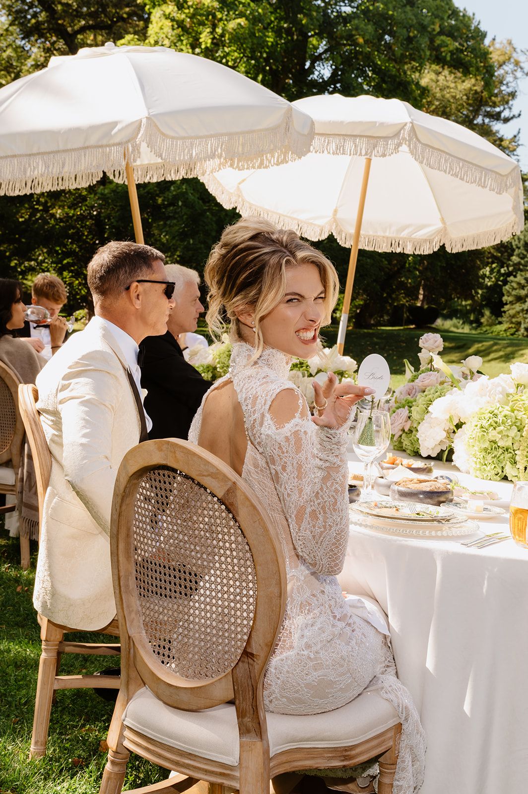 Elegant outdoor wedding reception with white parasols and rattan chairs in dappled sunlight.
