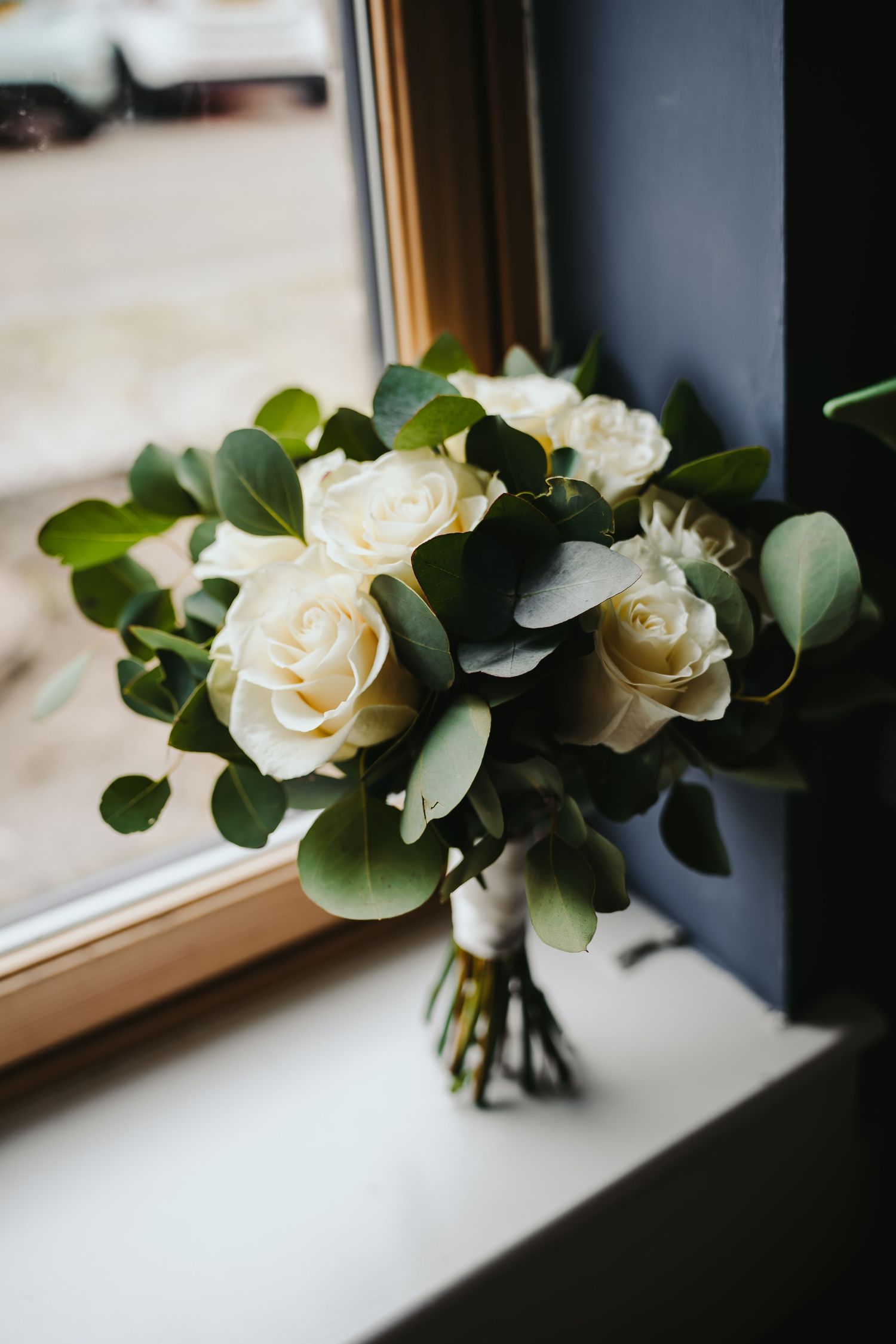 A delicate white rose bouquet with eucalyptus leaves sits elegantly on a windowsill in natural light.