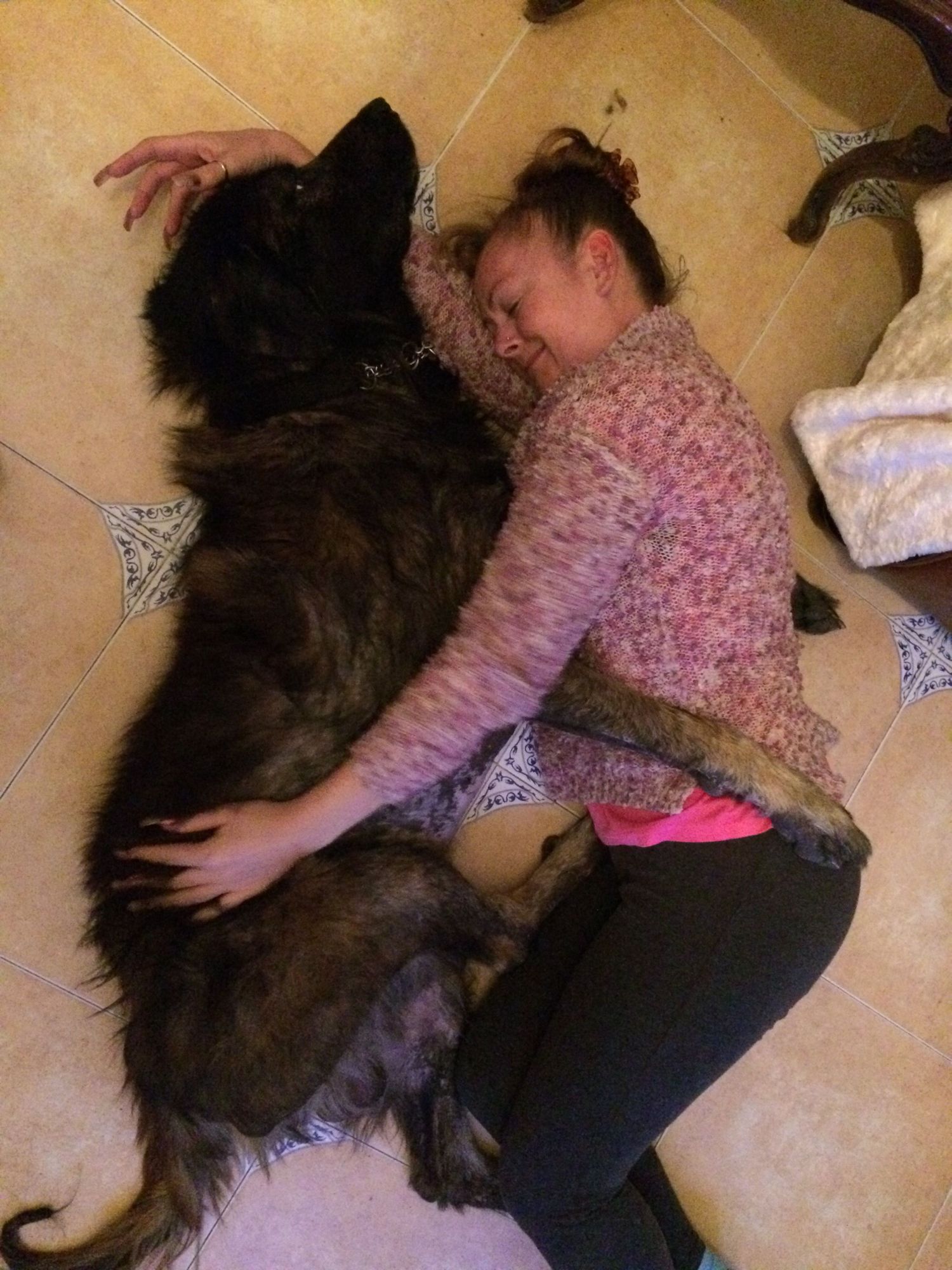 Young girl lying on tile floor cuddling and hugging a large fluffy black and tan dog.