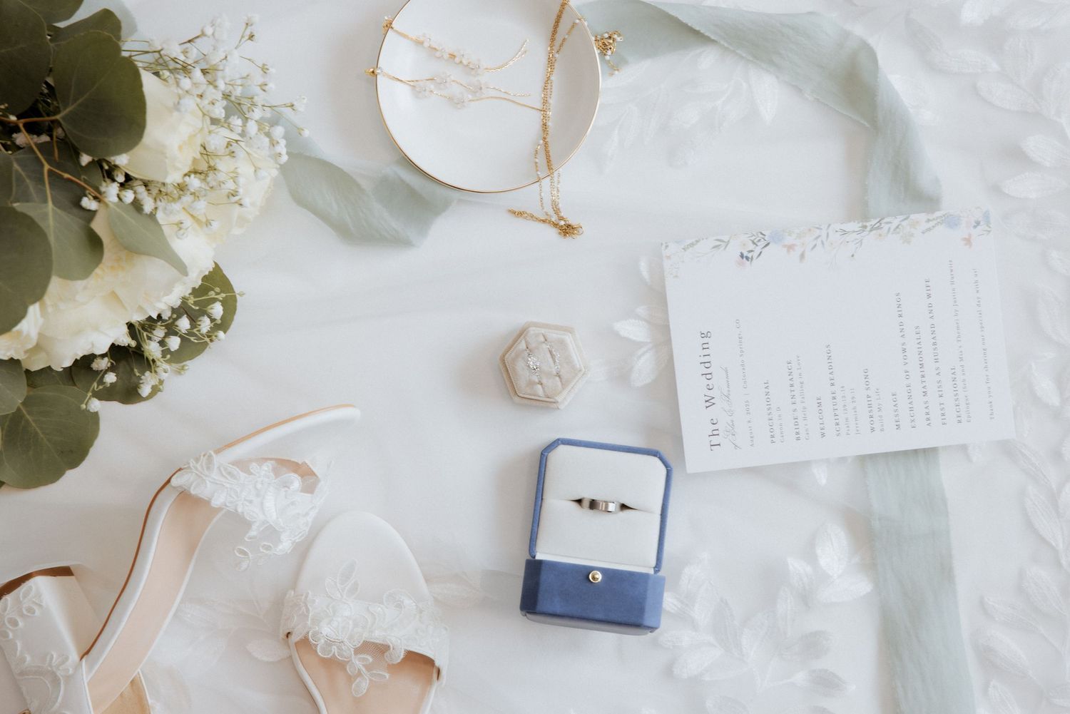 Wedding accessories including rings, floral arrangements, and delicate details arranged on white lace fabric.
