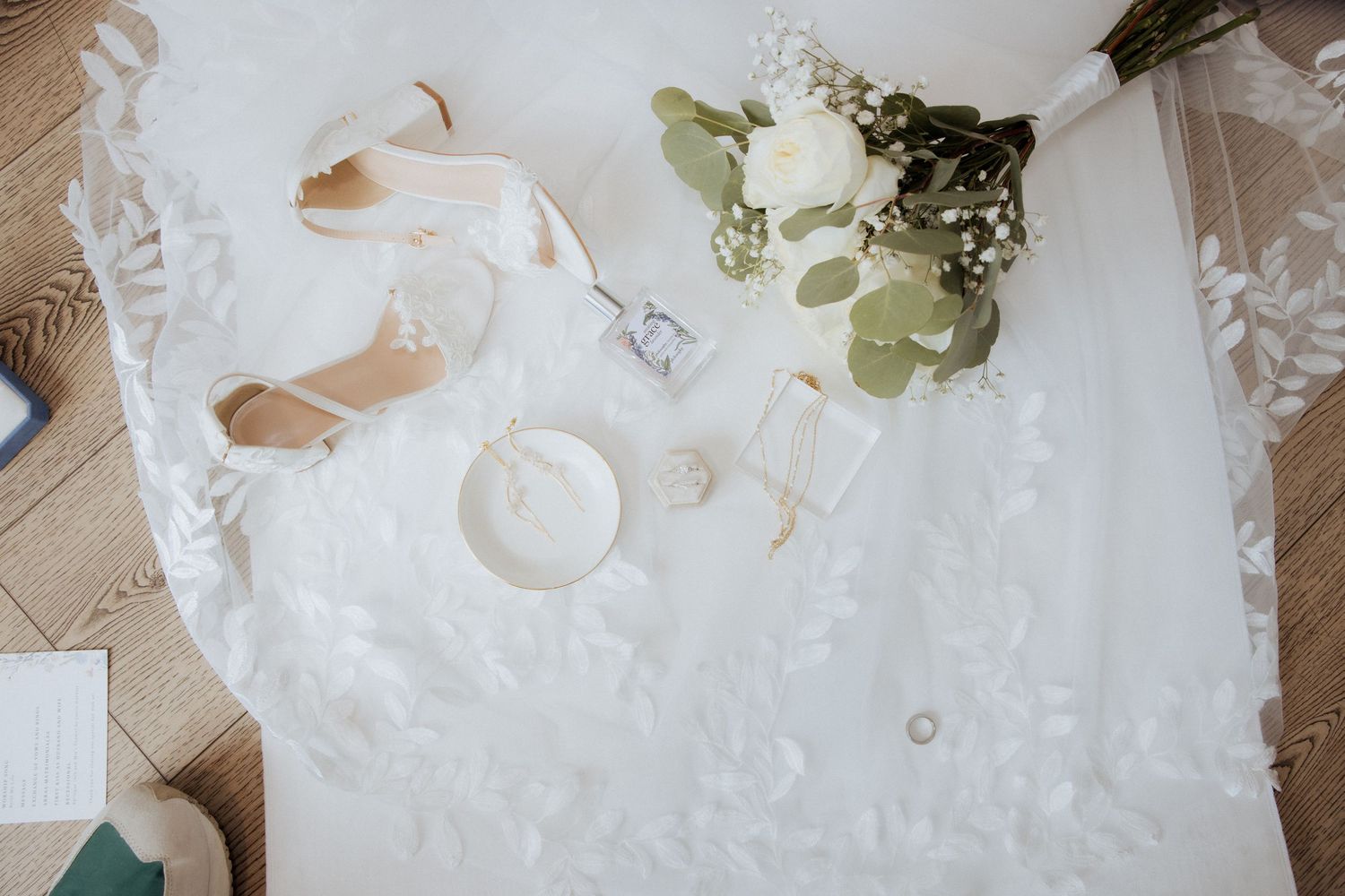 Wedding accessories including white dress, beige shoes, rings and eucalyptus bouquet arranged on light fabric.