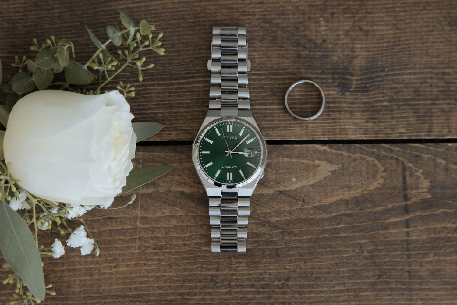 A silver wedding ring rests on a rustic wooden surface next to a stainless steel watch and white rose petals.