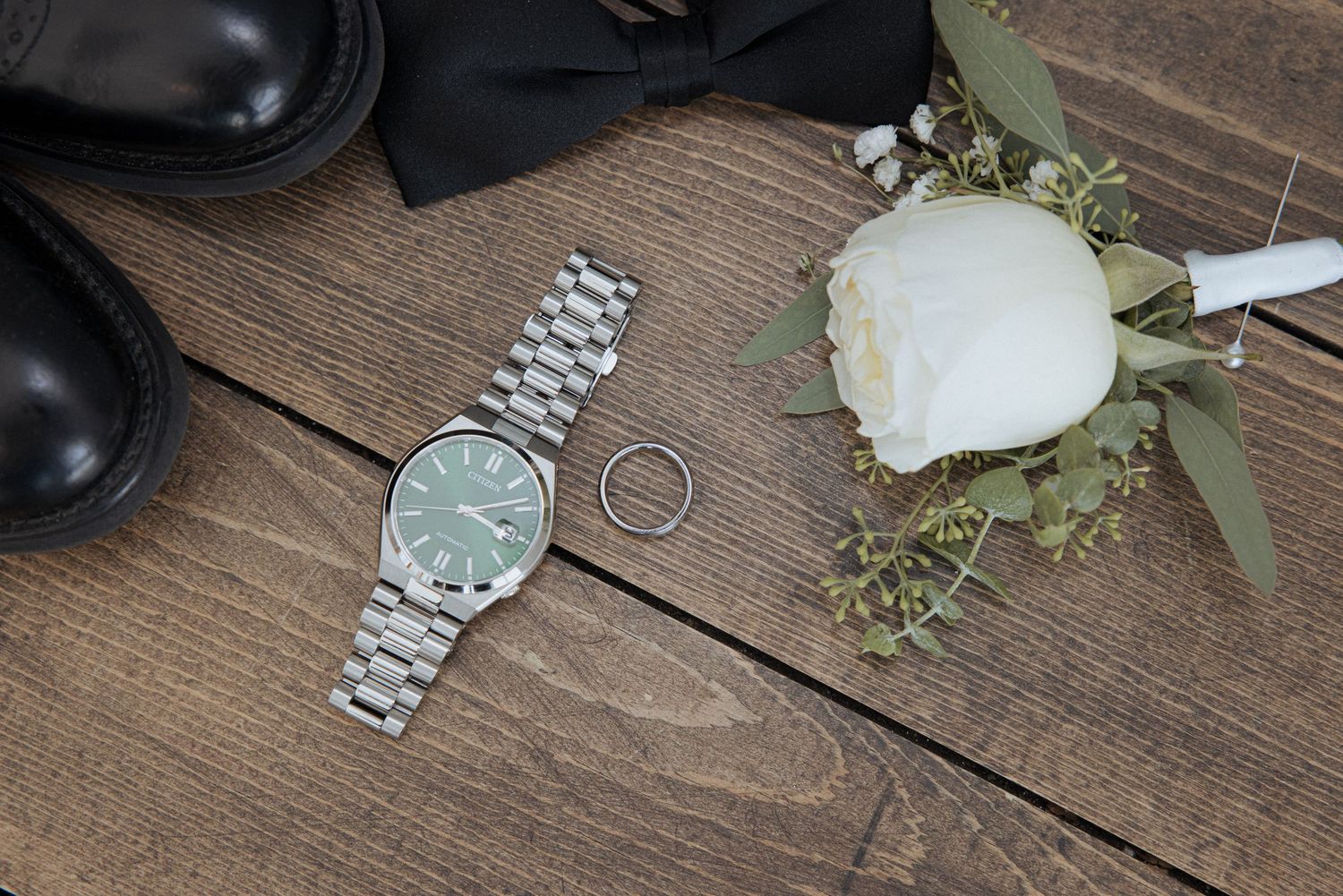 A silver wristwatch, wedding ring, and white rose with greenery arranged on a rustic wooden surface next to black shoes.