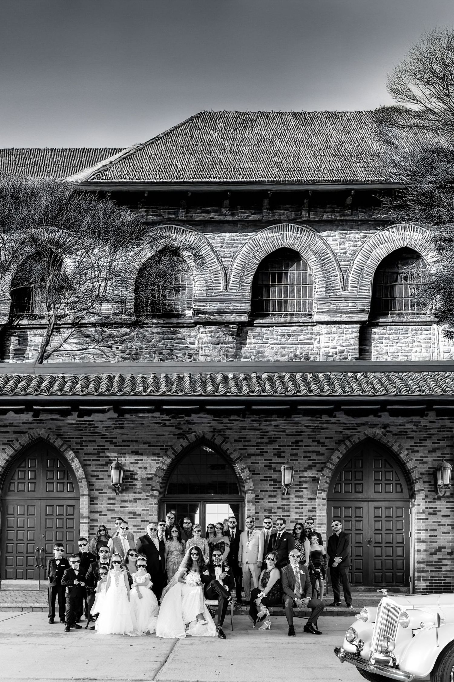 Black and white wedding photography sequence showing couple by vintage car outside Tudor-style building.