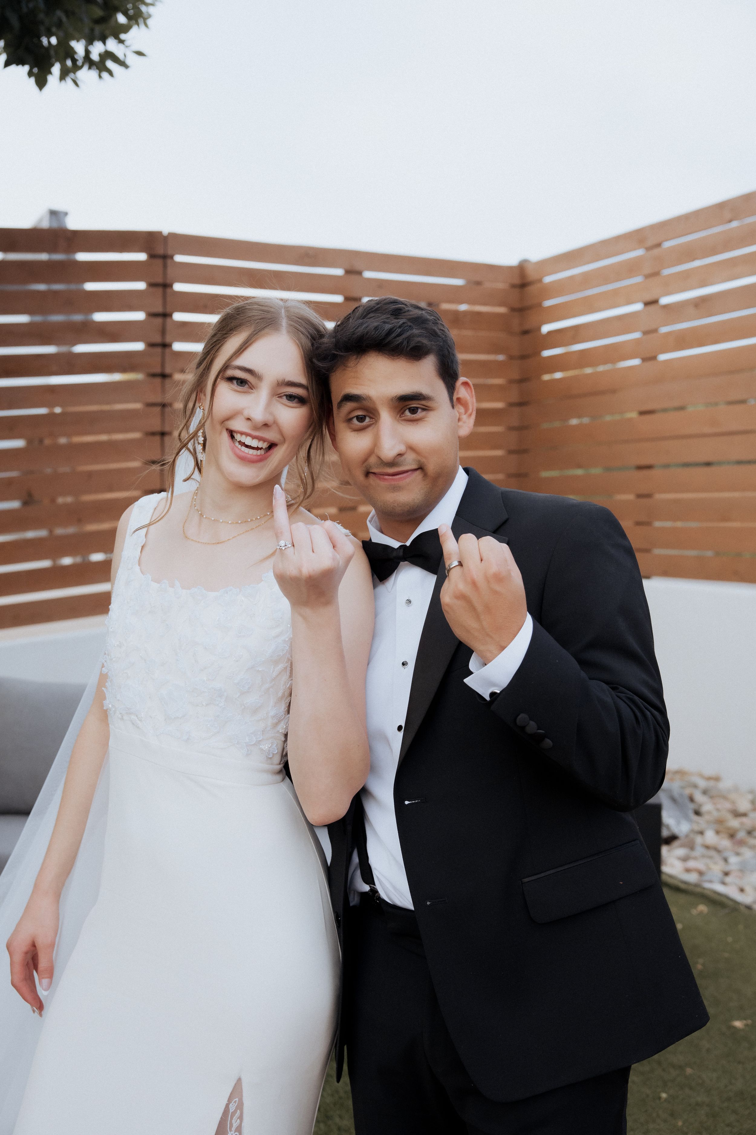 A couple shares joyful moments in formal wedding attire against a wooden fence backdrop.
