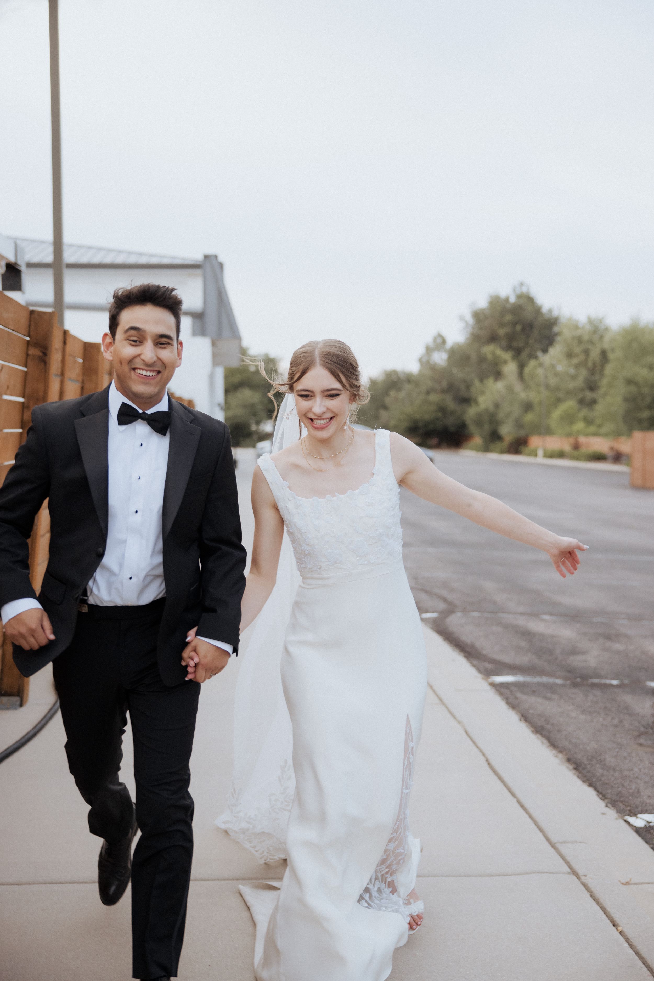 A couple in wedding attire running and laughing together on a sidewalk outside.