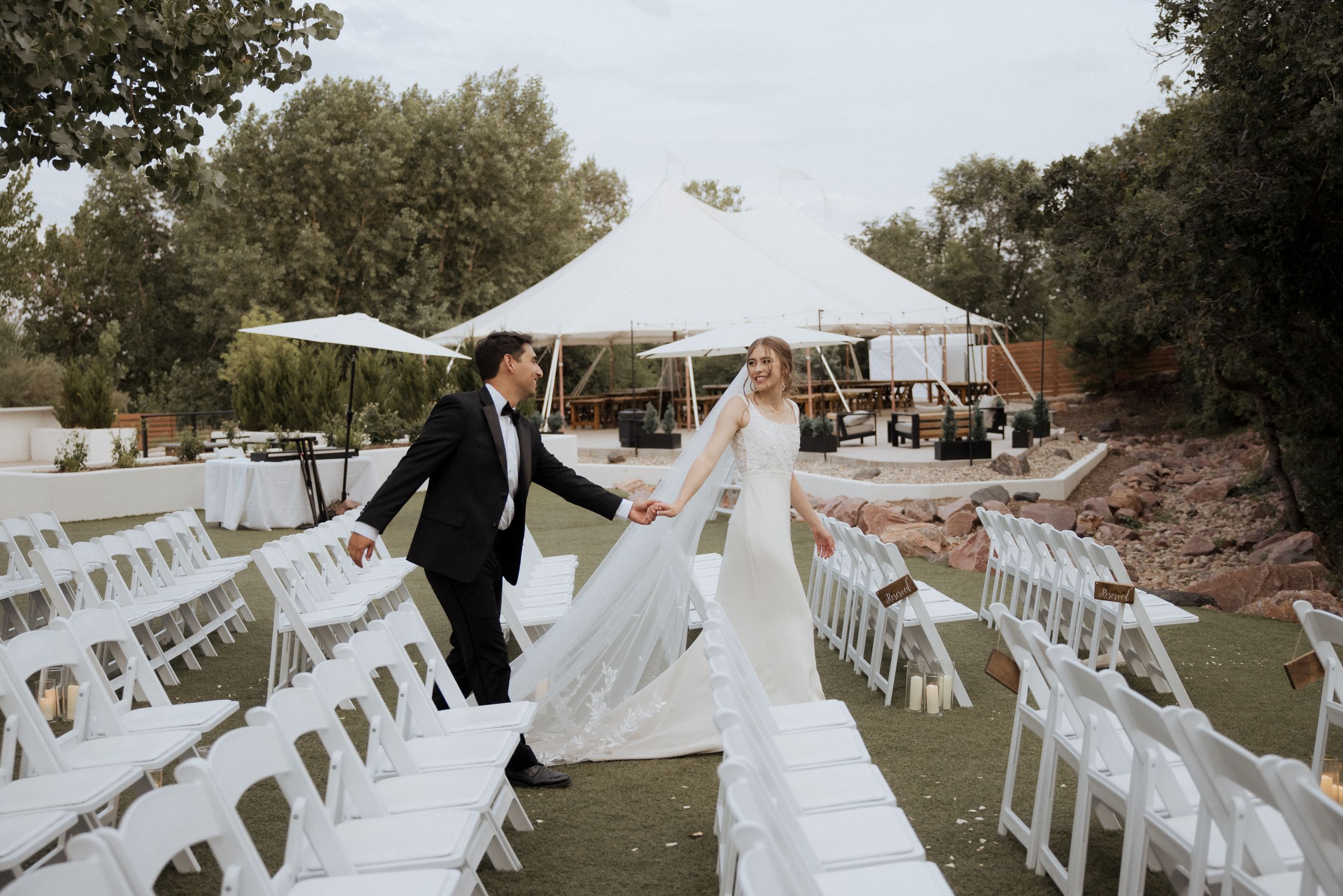 White folding chairs arranged in rows for an outdoor wedding ceremony under trees with a white tent in the distance.
