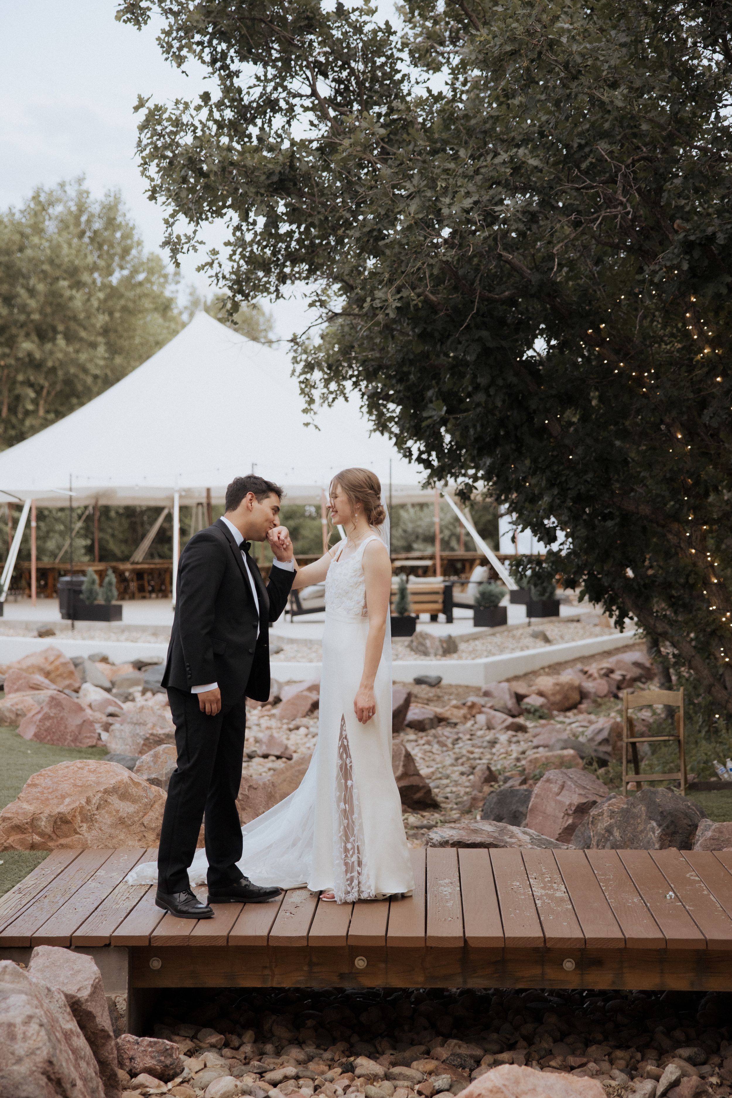 A couple shares a romantic moment on a wooden deck with a white wedding tent and trees in the background.