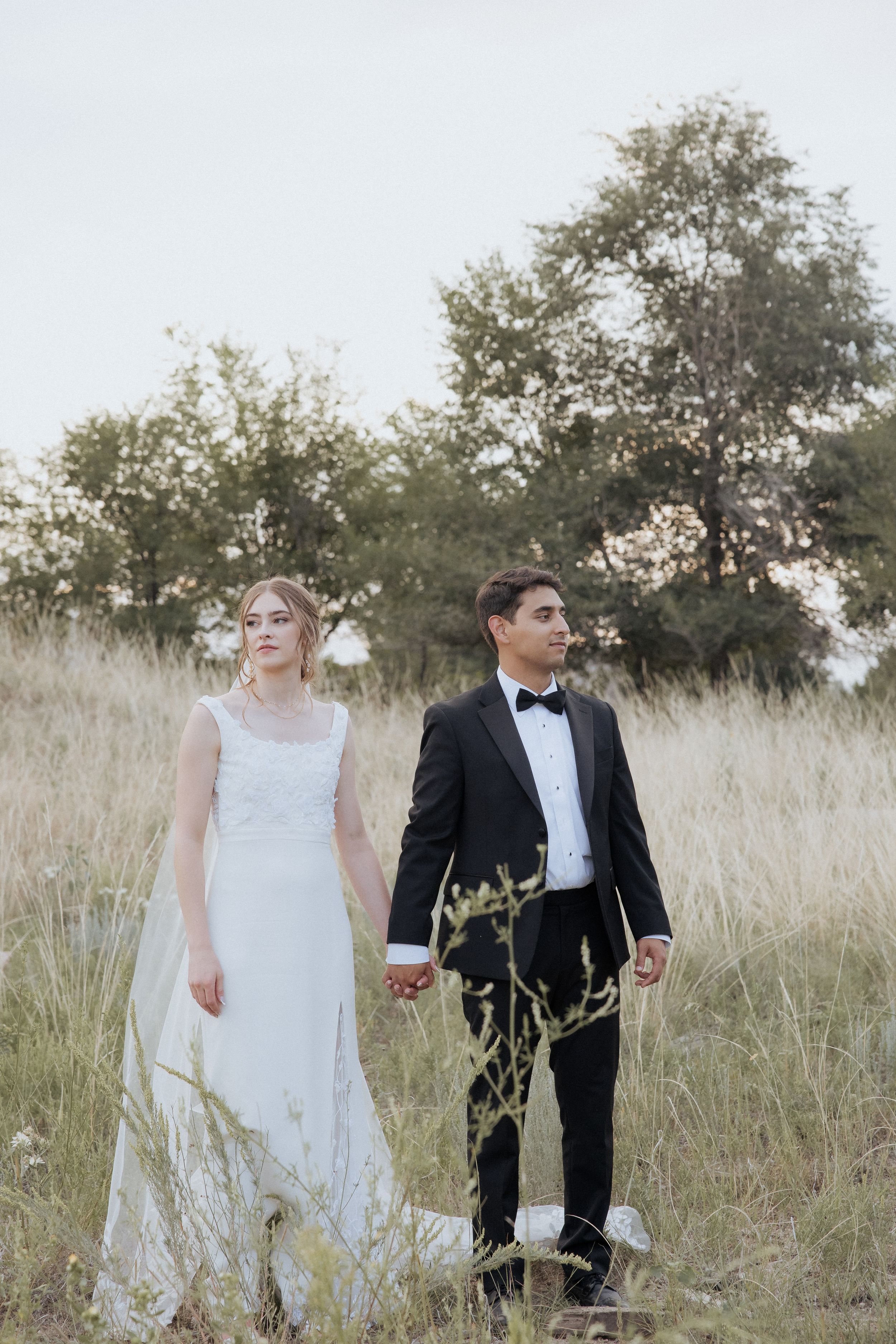 A couple in wedding attire stands together in a field of tall grass with trees in the background.