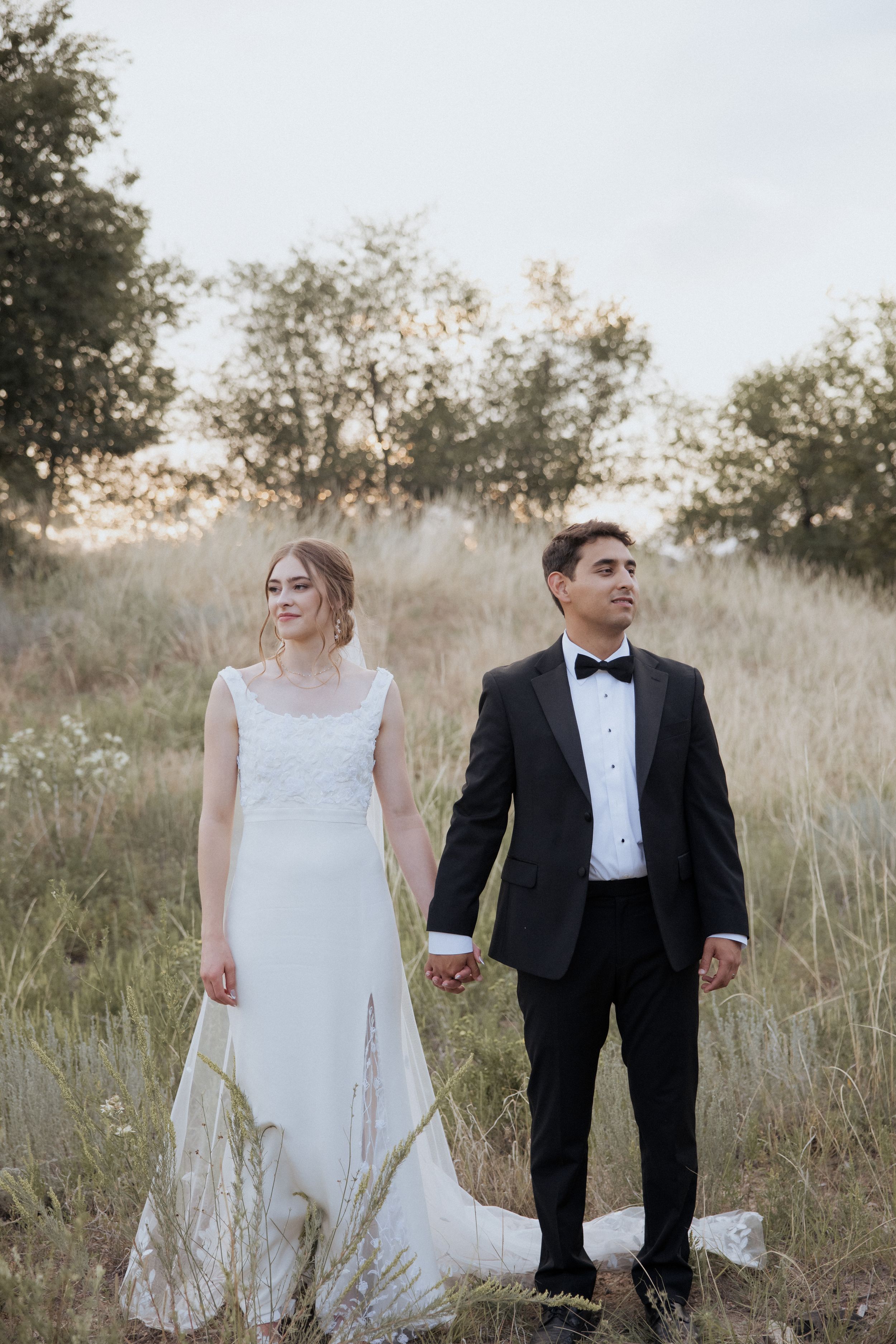 A couple in wedding attire sits together in a golden field at sunset holding wine glasses.