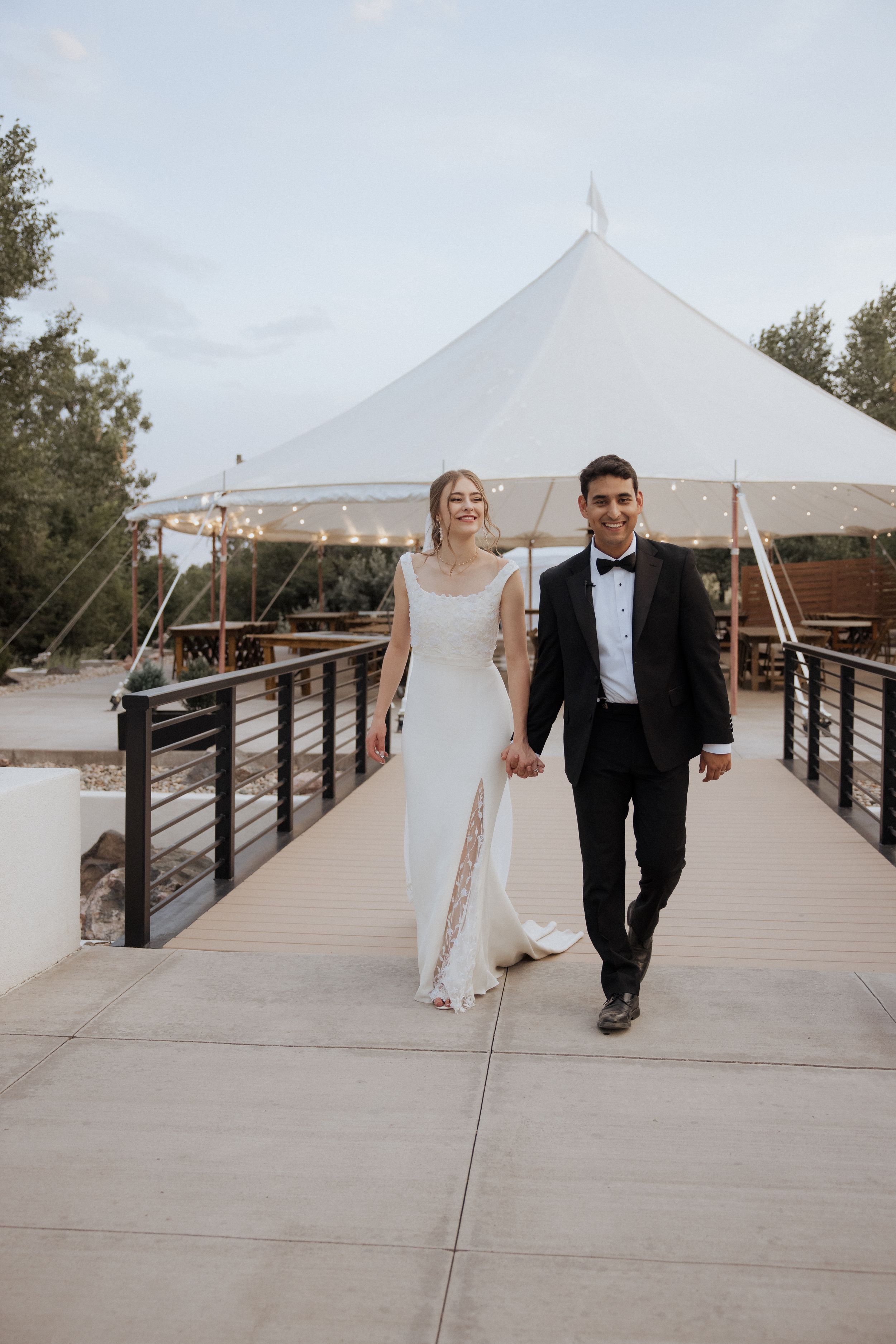 A newlywed couple walks along a wooden boardwalk with white event tents in the background.