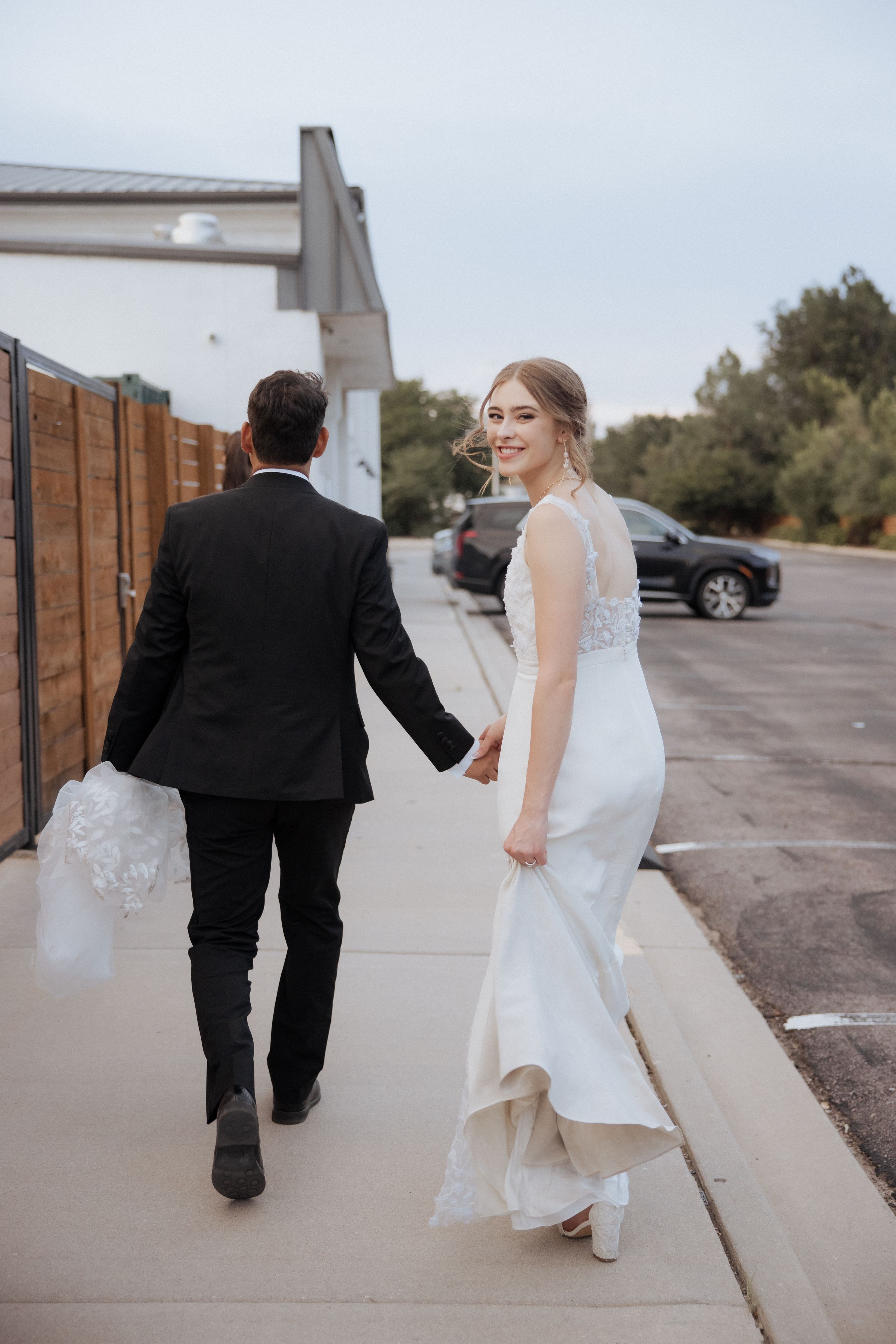 A couple walking together on a sidewalk, with the bride in a white gown and groom in a black suit.