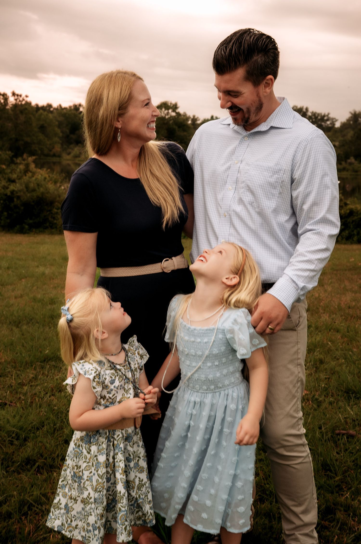 Happy family of four laughing together outdoors, parents with two young blonde girls in dresses on a green field.