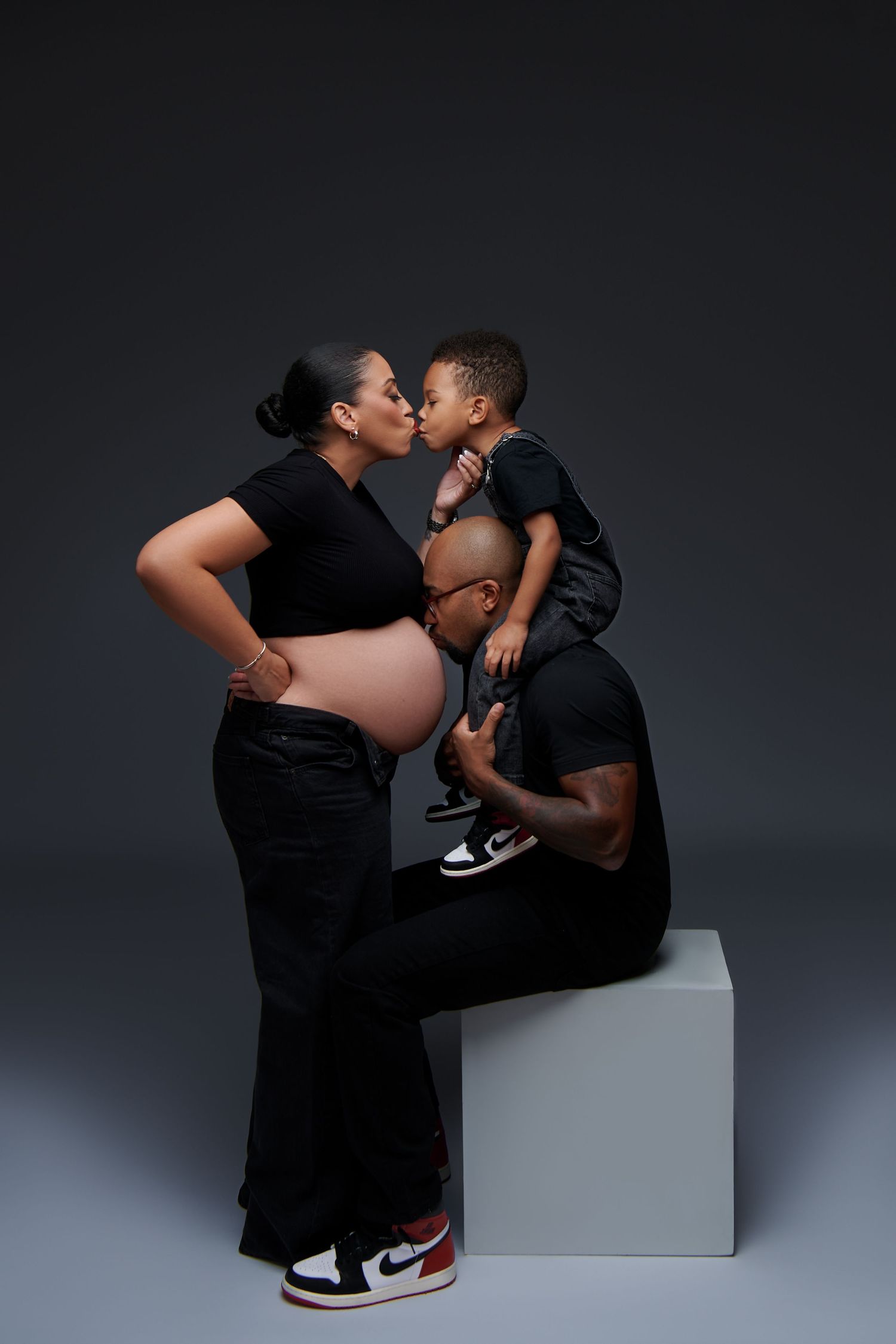A tender family moment captured in a studio portrait against a dark background, showing a pregnant woman in black.