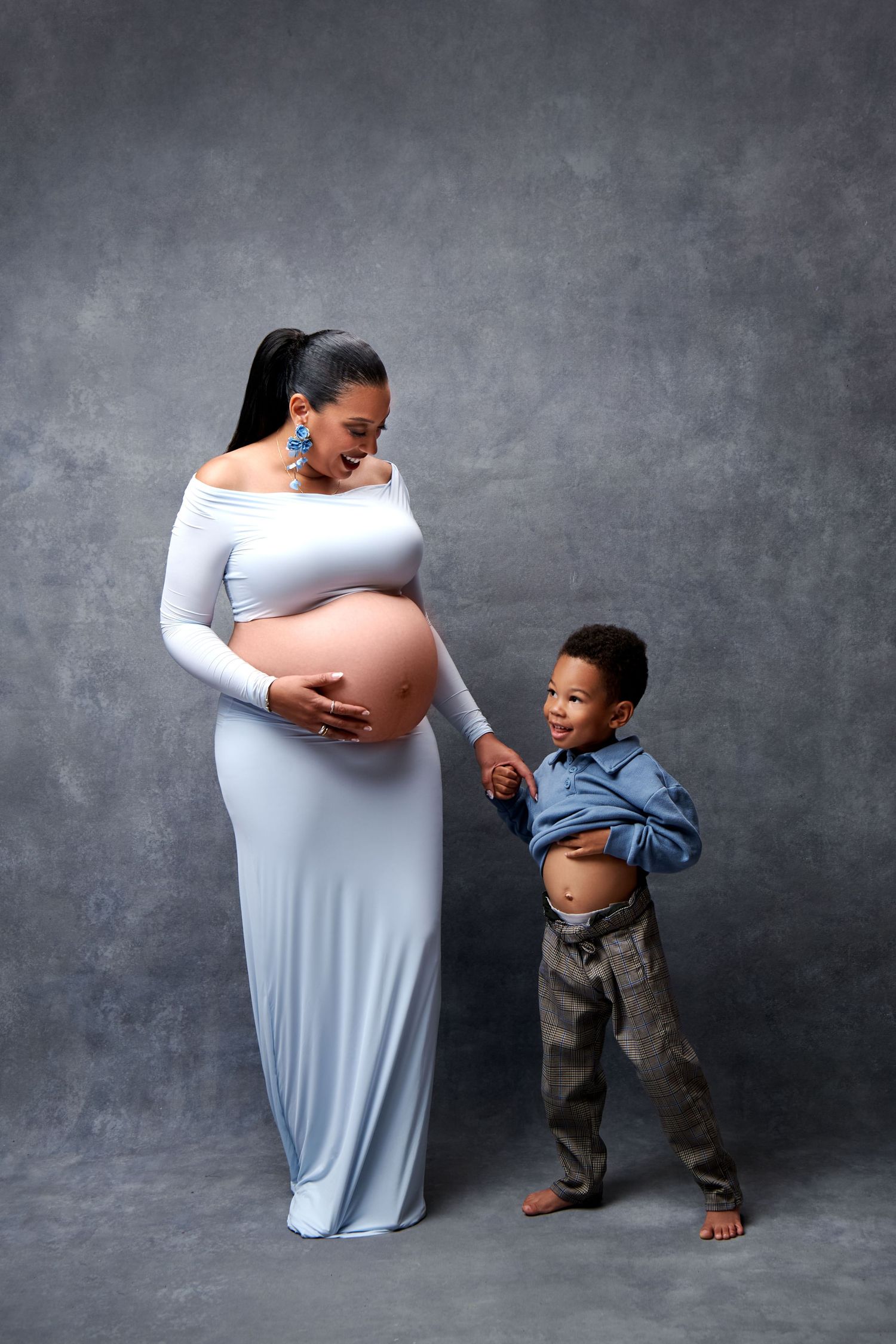 Maternity photoshoot showing interaction between small child and pregnant person against dark background.