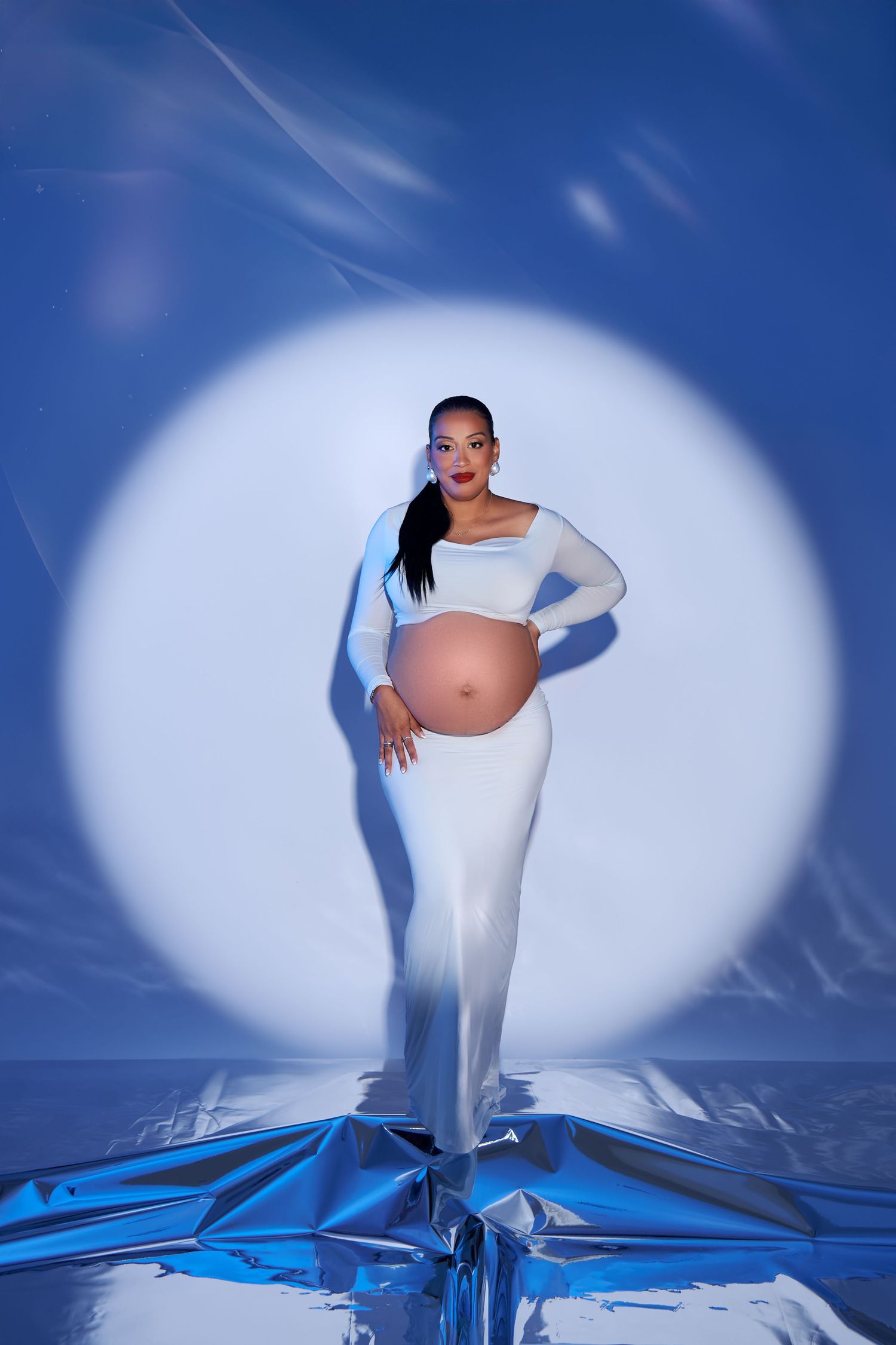 Artistic maternity photoshoot with white dress against blue background and dramatic spotlight effects.