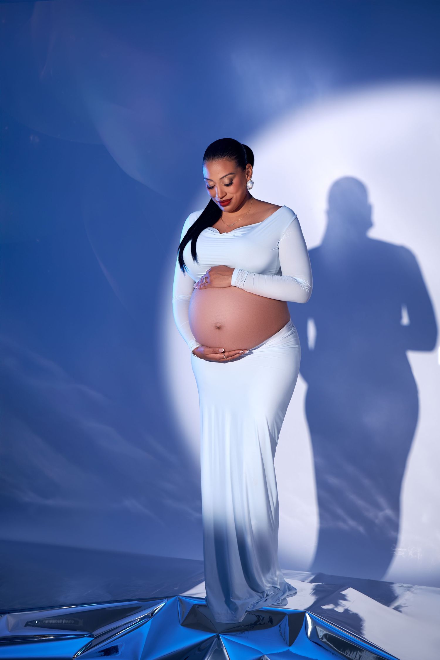 Expectant woman in flowing white dress poses against blue backdrop with dramatic lighting and shadow effects.