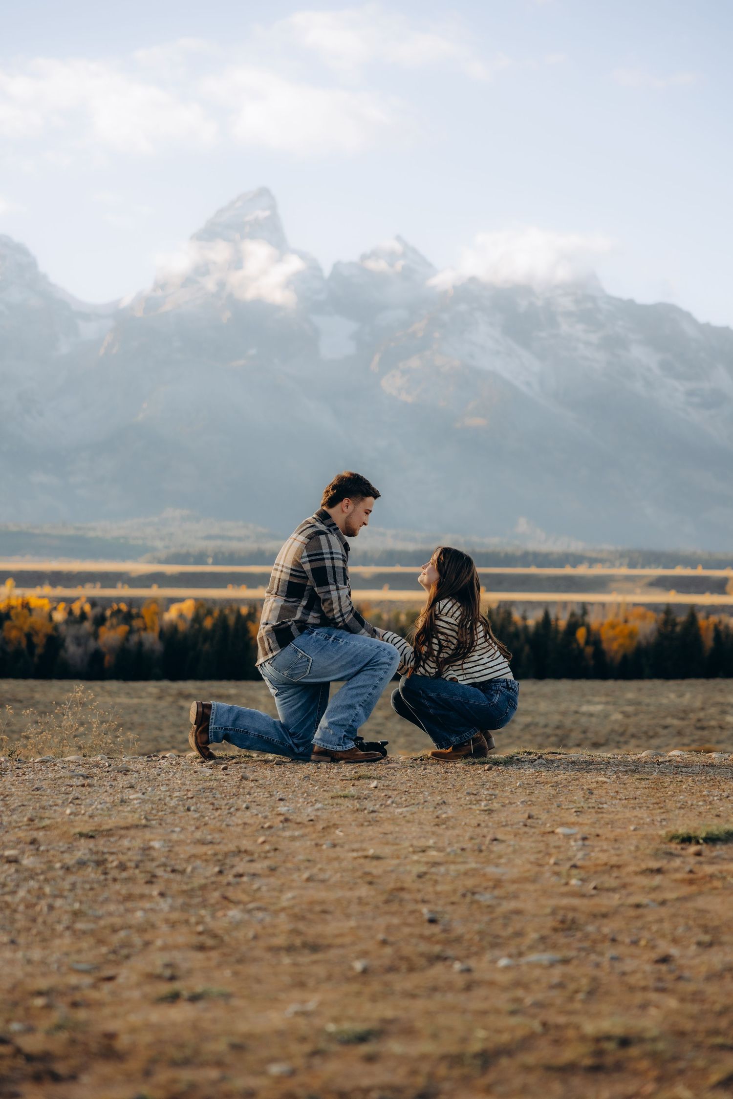 Two people sit together on a grassy field with majestic snow-capped mountains in the background at sunset.