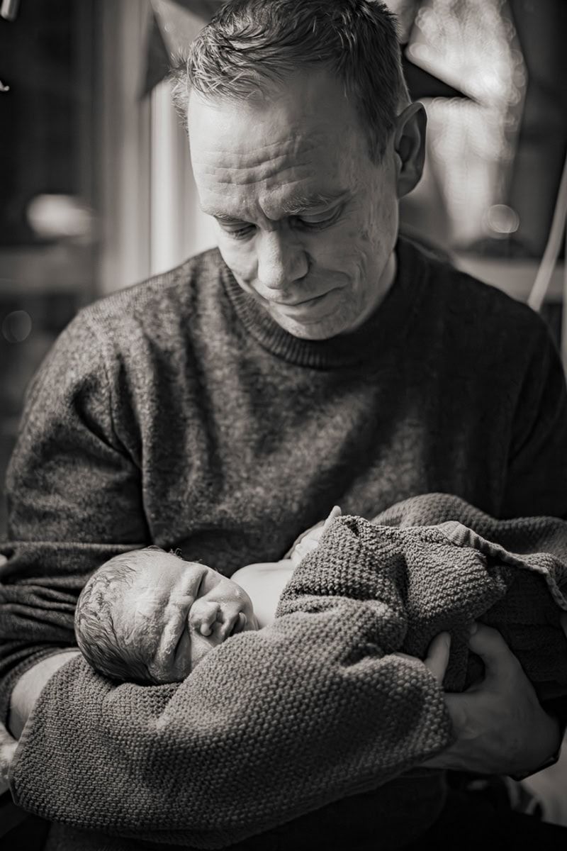Svartvitt förlossningsfoto av nybliven pappa som ser ner på sin nyfödda son