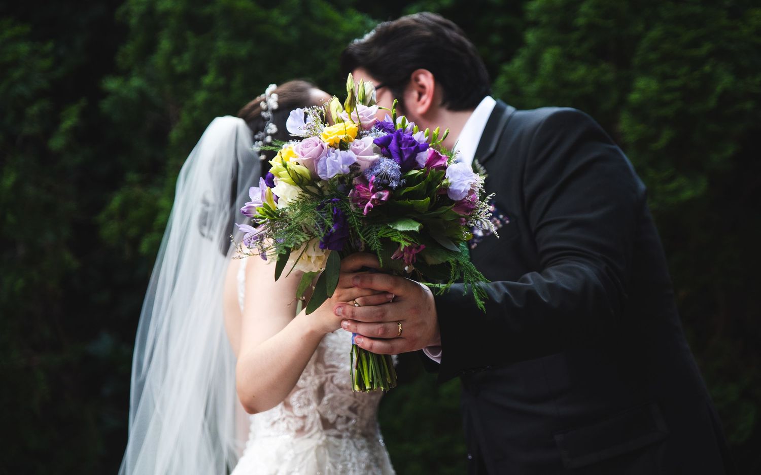 A newlywed couple kissing, hidden behind the bride's bouquet.