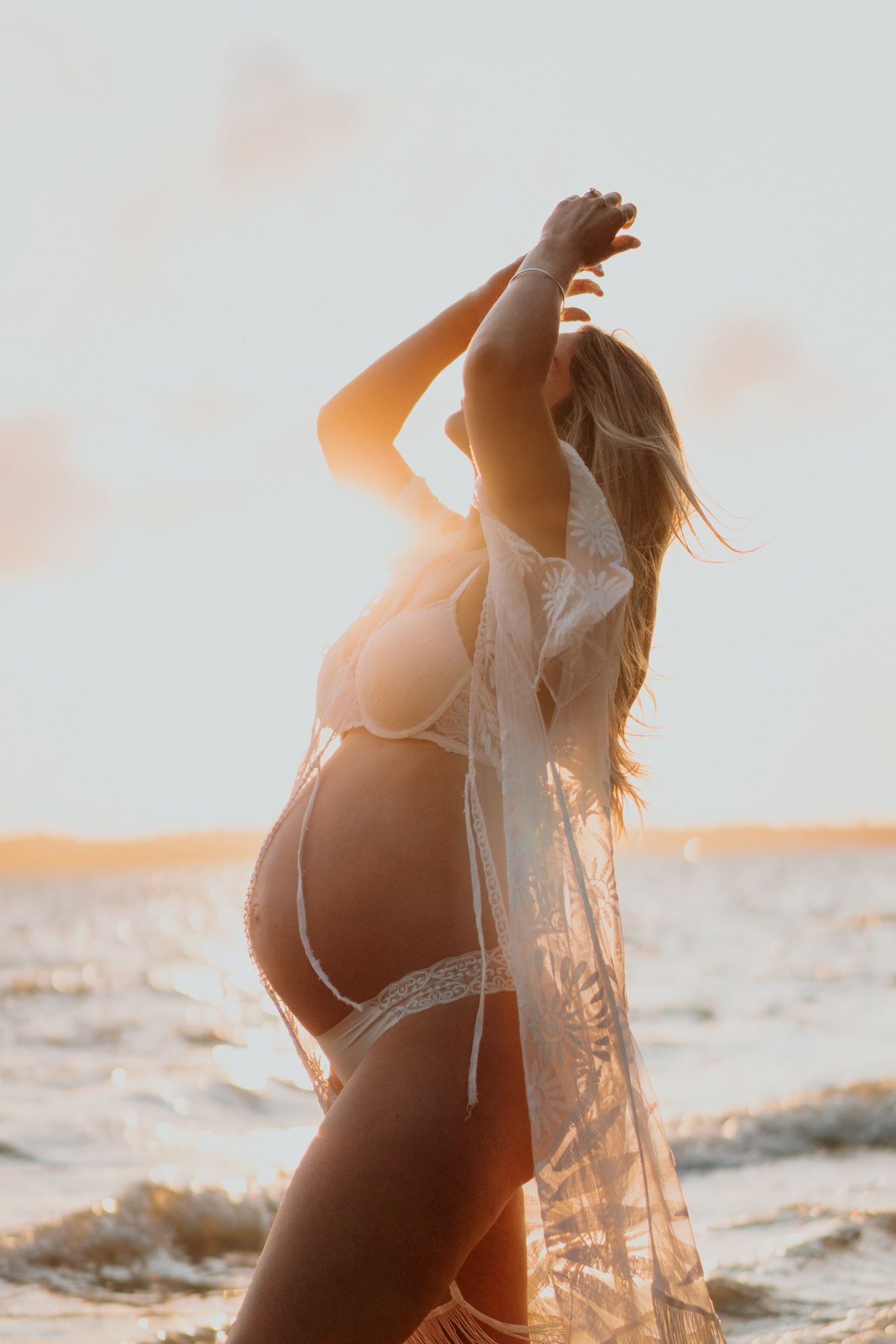 Silhouette of person in white lace dress standing on beach during golden sunset. Auckland maternity photographer