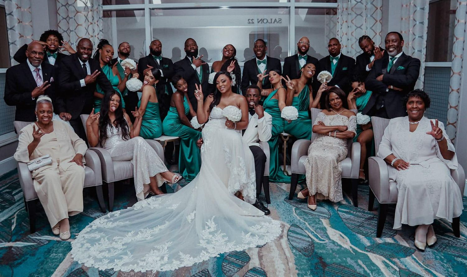 A large wedding party poses together in teal and white attire in an elegant venue.