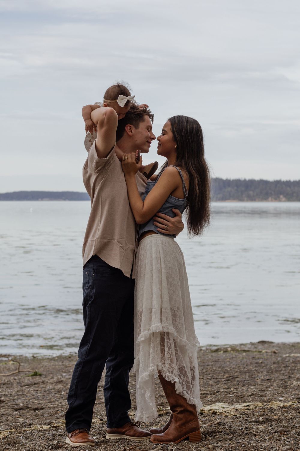 Couple nearly kissing on a rocky beach while man holds baby on shoulder, woman in white skirt and boots. taken by olympia photographer