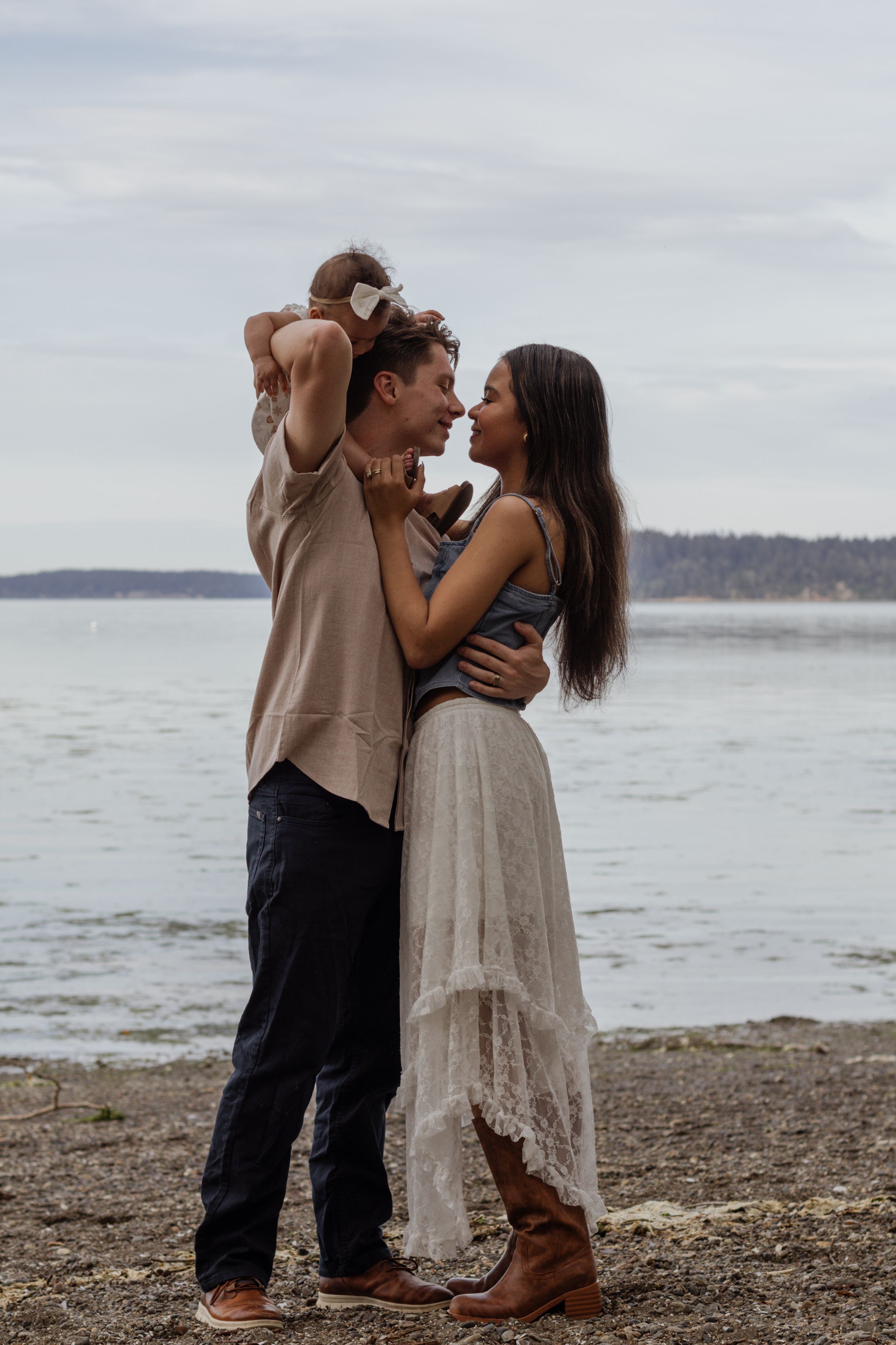Couple nearly kissing on a rocky beach while man holds baby on shoulder, woman in white skirt and boots. taken by olympia photographer