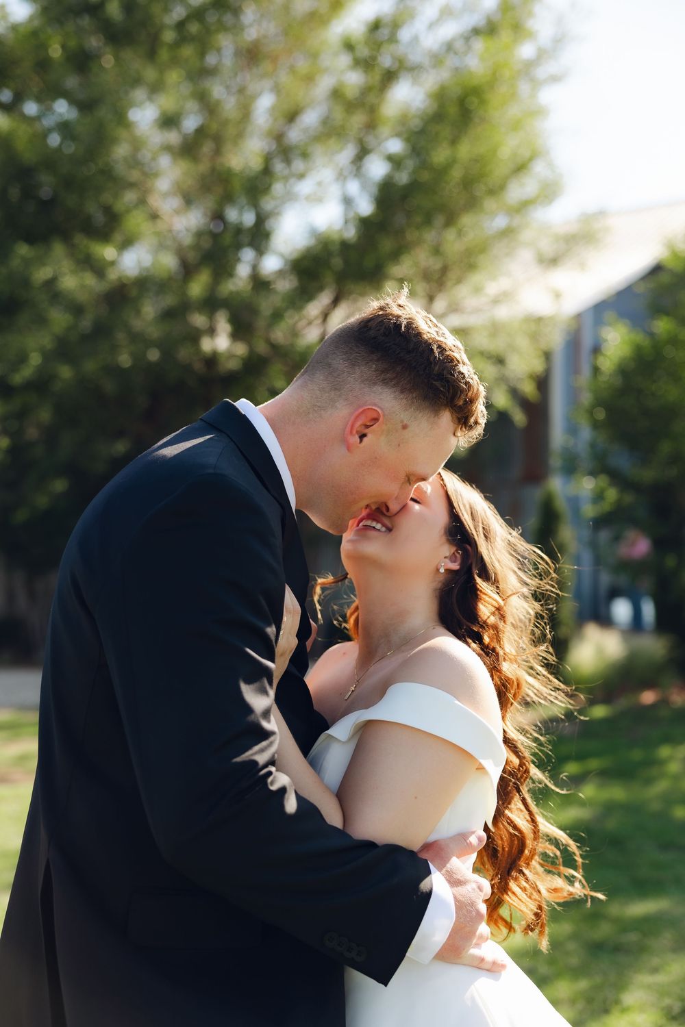 A couple in wedding attire share a romantic kiss outdoors on a sunny day with greenery in the background.