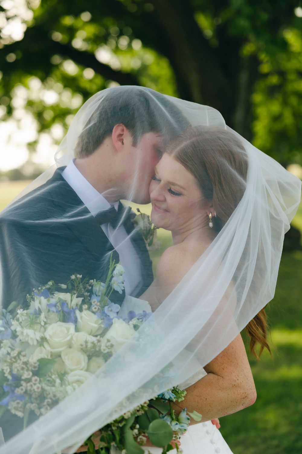 A romantic wedding couple shares a tender kiss outdoors while the bride's veil flows in the breeze with greenery in background.