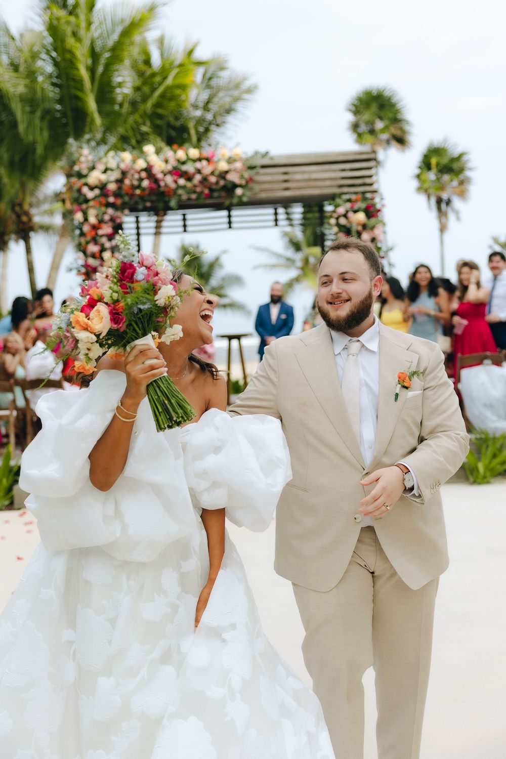 A joyful wedding couple shares candid moments on a tropical beach with palm trees and floral decor.